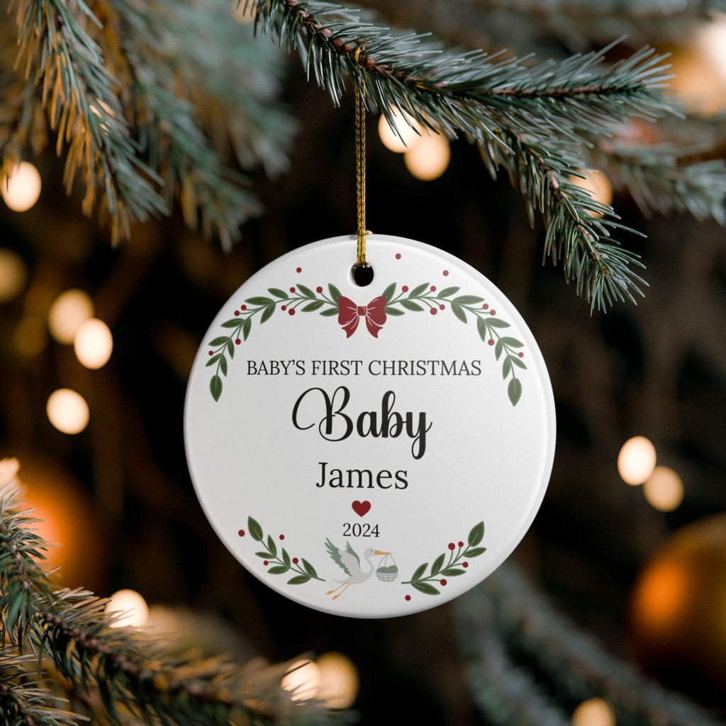 A personalized round white Christmas ornament hanging from a pine tree branch. The ornament features a gold hanging string and is printed with the text 'BABY'S FIRST CHRISTMAS Baby James 2024' along with green leafy borders and a small red bow.