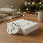 A round white Christmas ornament with personalized text and a green wreath design, resting on a white gift box on a wooden table with a blurred holiday background.