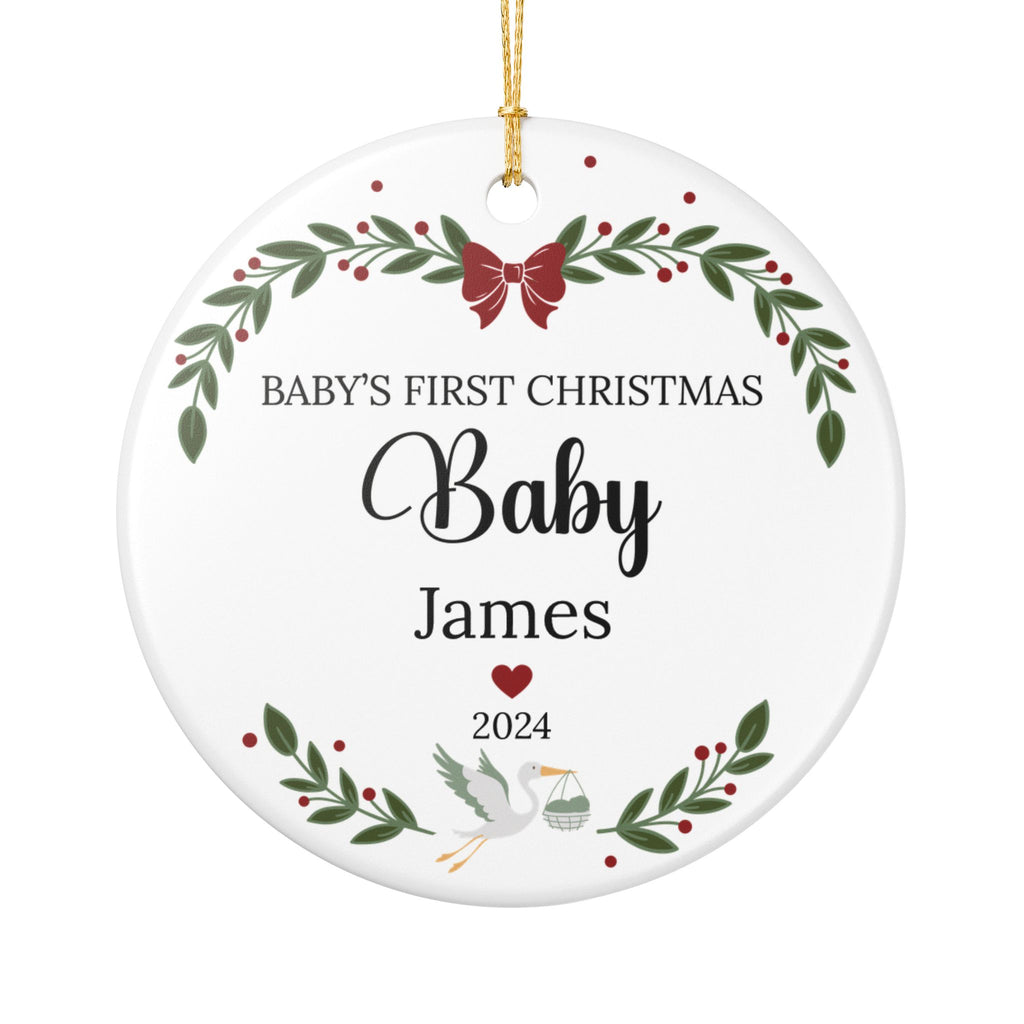 Round white ceramic Christmas ornament with personalized text reading 'Baby's First Christmas Baby James 2024,' decorated with green holly leaves, red berries, a red bow, a white stork, and a gold hanging string.