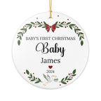 Round white ceramic Christmas ornament with personalized text reading 'Baby's First Christmas Baby James 2024,' decorated with green holly leaves, red berries, a red bow, a white stork, and a gold hanging string.