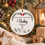 Personalized round white Christmas ornament hanging from a gold string in a festive tree setting, with printed text reading 'Baby's First Christmas Baby James 2024' and holiday illustrations including a stork, green leaves, red berries, and a red bow.