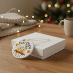 A round white ceramic Halloween ornament with a 'Boo Crew' design, hanging from a gold string next to a white gift box on a wooden table with a blurred Christmas tree and cozy background.