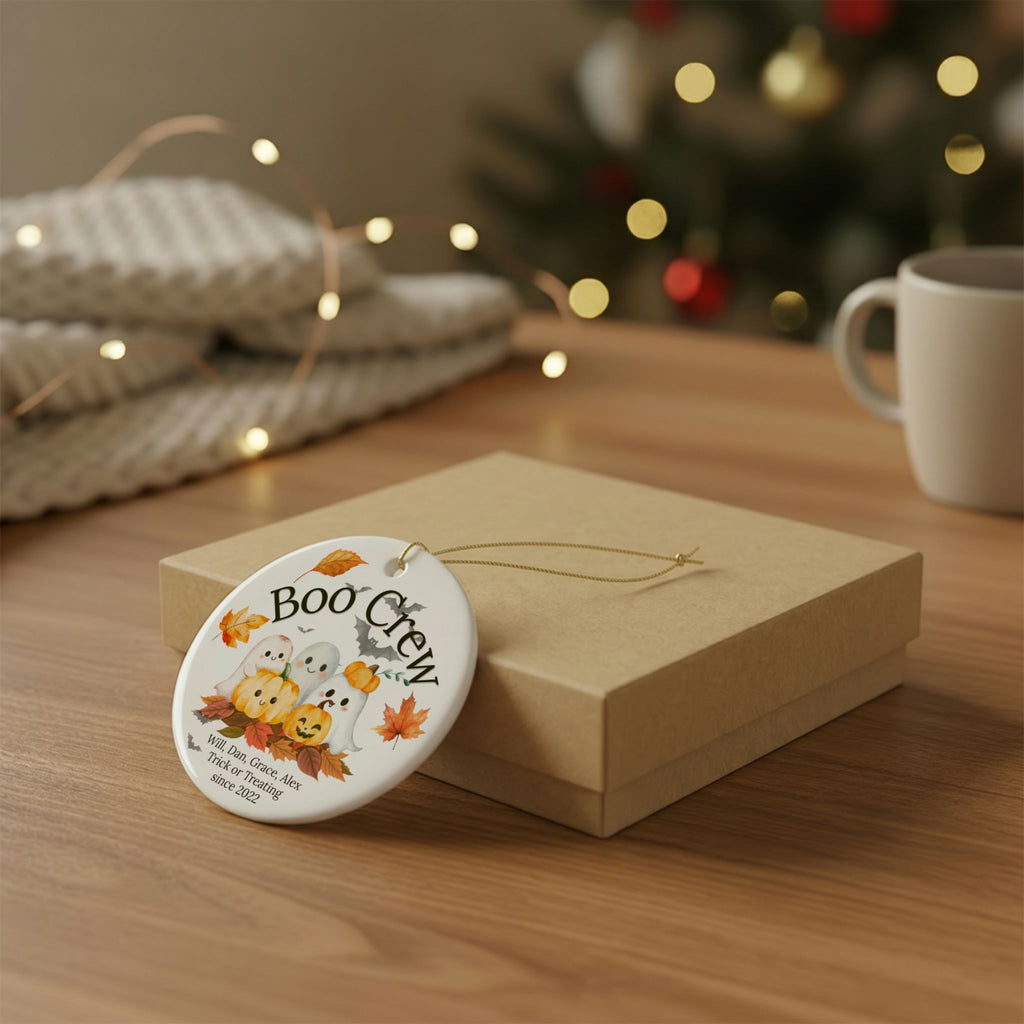 A round white ceramic Halloween ornament with a 'Boo Crew' design, resting on a brown gift box on a wooden table with a cozy, blurred holiday background.