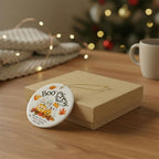 A round white ceramic Halloween ornament with a 'Boo Crew' design, resting on a brown gift box on a wooden table with a cozy, blurred holiday background.