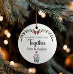 A round white ceramic Christmas ornament hanging from a pine branch. The ornament is printed with personalized holiday text reading 'OUR FIRST CHRISTMAS Together Alex & Bailey 2025' with a red bow, green leaves, and small cactus illustrations.