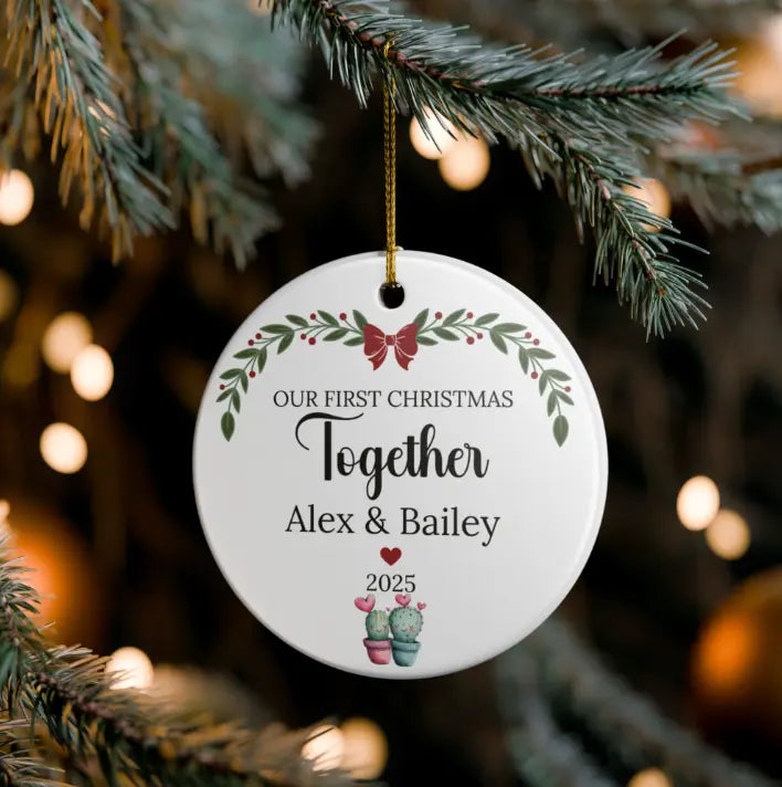 A round white ceramic Christmas ornament hanging from a pine branch. The ornament is printed with personalized holiday text reading 'OUR FIRST CHRISTMAS Together Alex & Bailey 2025' with a red bow, green leaves, and small cactus illustrations.