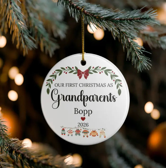 A round white personalized Christmas ornament hanging from a pine branch. The ornament features a gold hanging string and printed text reading 'OUR FIRST CHRISTMAS AS Grandparents Bopp 2026' with green leaves, a red bow, and small holiday figures at the bottom.