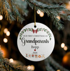 A round white personalized Christmas ornament hanging from a pine branch. The ornament features a gold hanging string and printed text reading 'OUR FIRST CHRISTMAS AS Grandparents Bopp 2026' with green leaves, a red bow, and small holiday figures at the bottom.