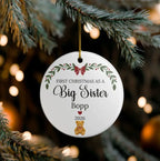 A round white Christmas ornament hanging from a pine branch, printed with the message 'FIRST CHRISTMAS AS A Big Sister Bopp 2026' and decorated with green leaves, red berries, a red bow, and a small teddy bear.