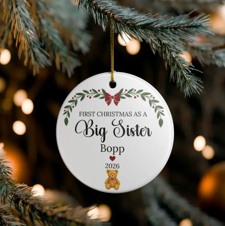 A round white Christmas ornament hanging from a pine branch, printed with the message 'FIRST CHRISTMAS AS A Big Sister Bopp 2026' and decorated with green leaves, red berries, a red bow, and a small teddy bear.