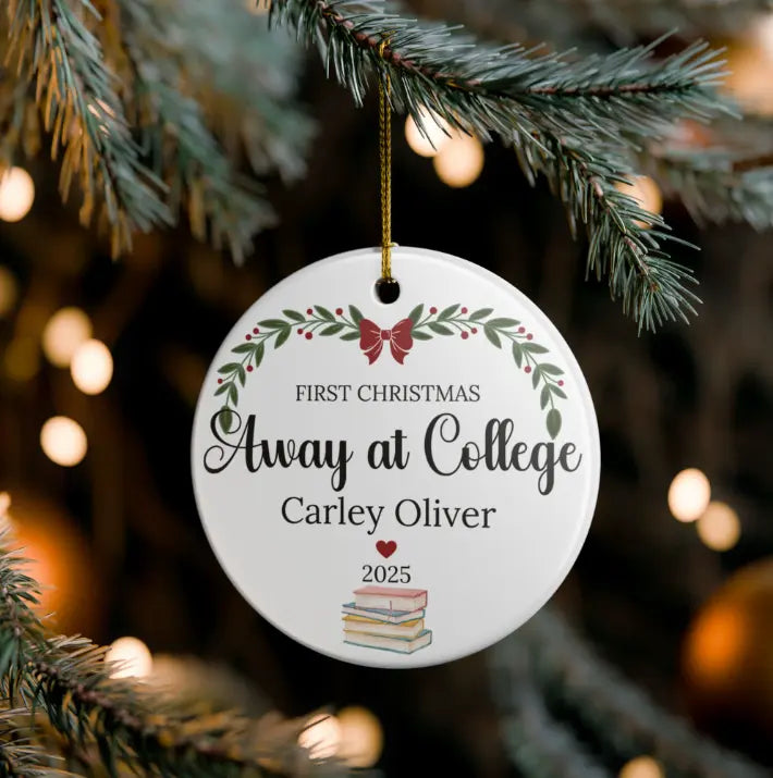 A personalized round white Christmas ornament hanging from a pine tree branch. The ornament features a gold hanging string, a red bow, green leafy branches, and the text 'FIRST CHRISTMAS Away at College Carley Oliver 2025' along with a small stack of books illustration.