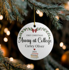 A personalized round white Christmas ornament hanging from a pine tree branch. The ornament features a gold hanging string, a red bow, green leafy branches, and the text 'FIRST CHRISTMAS Away at College Carley Oliver 2025' along with a small stack of books illustration.