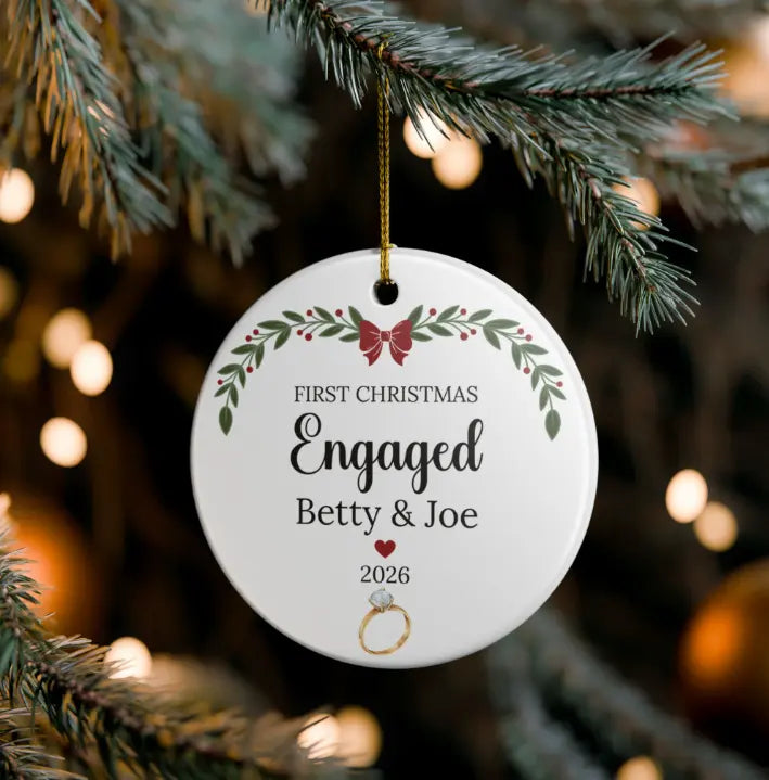 A round white Christmas ornament hanging from a pine branch, printed with the words “First Christmas Engaged Betty & Joe 2026” and a small ring illustration, with blurred holiday lights in the background.