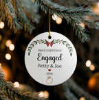 A round white Christmas ornament hanging from a pine branch, printed with the words “First Christmas Engaged Betty & Joe 2026” and a small ring illustration, with blurred holiday lights in the background.
