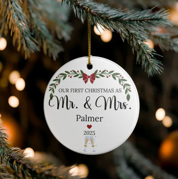 A personalized round white ceramic Christmas ornament hanging from a pine tree branch with warm bokeh lights in the background. The ornament features a green holly leaf border, a red bow, and the text 'OUR FIRST CHRISTMAS AS Mr.