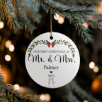 A personalized round white ceramic Christmas ornament hanging from a pine tree branch with warm bokeh lights in the background. The ornament features a green holly leaf border, a red bow, and the text 'OUR FIRST CHRISTMAS AS Mr.