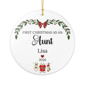 A round white personalized Christmas ornament with a gold hanging string. The ornament features printed black text reading 'FIRST CHRISTMAS AS AN Aunt' with the name 'Lisa' below, plus holiday illustrations including a green wreath with red berries, a red bow, a red heart, and Christmas stockings on a clothesline.