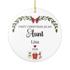 A round white personalized Christmas ornament with a gold hanging string. The ornament features printed black text reading 'FIRST CHRISTMAS AS AN Aunt' with the name 'Lisa' below, plus holiday illustrations including a green wreath with red berries, a red bow, a red heart, and Christmas stockings on a clothesline.