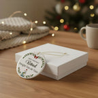 A round Christmas ornament with a colorful pastel border and a pink unicorn illustration hangs from a gold string, resting on a white gift box on a wooden table. The background is softly blurred with warm fairy lights, a knitted blanket, and a mug, creating a cozy holiday atmosphere.