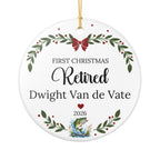 A round white Christmas ornament with a gold hanging string, featuring personalized text that reads 'FIRST CHRISTMAS Retired Dwight Van de Vate 2026' with green leafy garlands, red berries, a red bow, and a small fish illustration near the bottom.