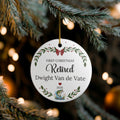 A personalized round white Christmas ornament hanging from a pine tree branch. The ornament features a gold hanging string and printed text reading 'FIRST CHRISTMAS Retired Dwight Van de Vate 2026' with green and red floral illustrations.